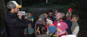 Eine Gruppe von Kindern steht in einem Wald, einige tragen Mützen und Jacken, und ein Kind hält eine Taschenlampe.