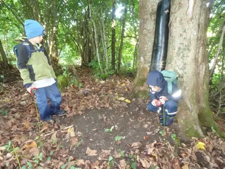 Zwei Kinder in einem Wald untersuchen einen Baumstamm. Eines kniet mit einem Kompass, und das andere hält einen Kompass.