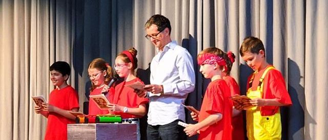 Eine Gruppe von Kindern in roten Hemden tritt auf der Bühne auf, mit einem Mann in einem weißen Hemd und einer Brille vor ihnen. Sie halten ein Buch und ein Mikrofon.