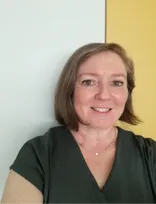 Eine Frau mit kurzen braunen Haaren lächelt und trägt ein dunkelgrünes Hemd. Sie hat eine Halskette mit einem Anhänger und steht vor einer gelben Wand.