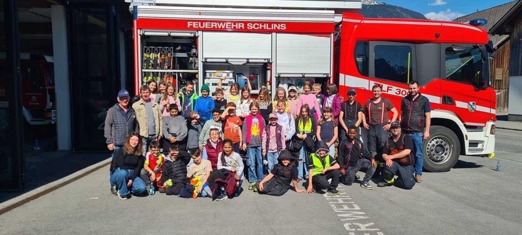 Eine Gruppe von Kindern und Erwachsenen posiert vor einem Feuerwehrauto mit der Aufschrift Feuerwehr Schins LF 30. Der Ort befindet sich in der Nähe eines Gebäudes und Berge unter einem teilweise bewölkten Himmel.