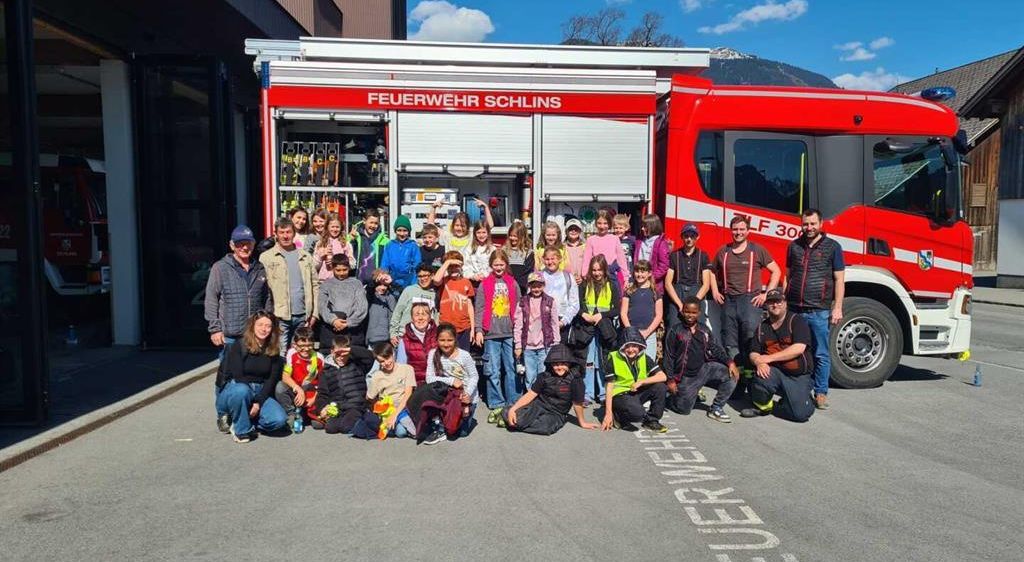 Eine Gruppe von Kindern und Erwachsenen posiert vor einem Feuerwehrauto mit der Aufschrift Feuerwehr Schins LF 30. Der Ort befindet sich in der Nähe eines Gebäudes und Berge unter einem teilweise bewölkten Himmel.