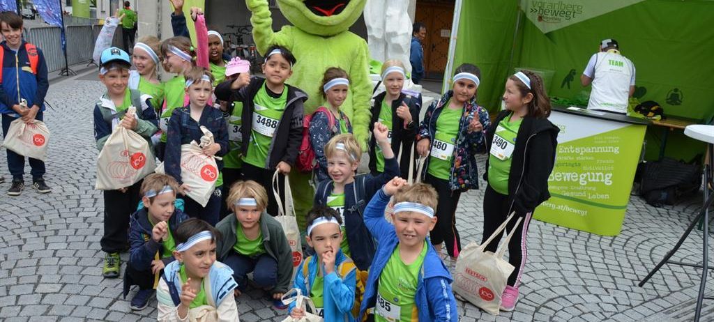 Eine Gruppe von Kindern in grünen Shirts und Stirnbändern posiert für ein Foto mit einem großen grünen Frosch-Maskottchen vor einem grünen Zelt.