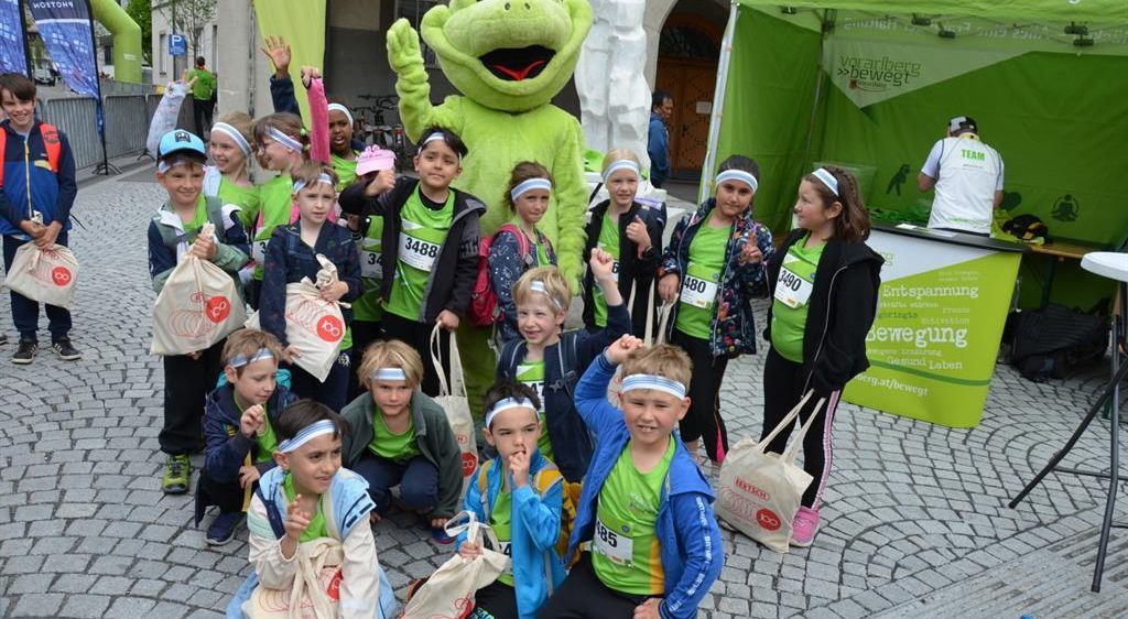 Eine Gruppe von Kindern in grünen Shirts und Stirnbändern posiert für ein Foto mit einem großen grünen Frosch-Maskottchen vor einem grünen Zelt.
