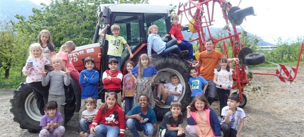 Eine Gruppe von Kindern und Erwachsenen posiert für ein Foto vor einem Traktor, mit Bäumen und Bergen im Hintergrund.