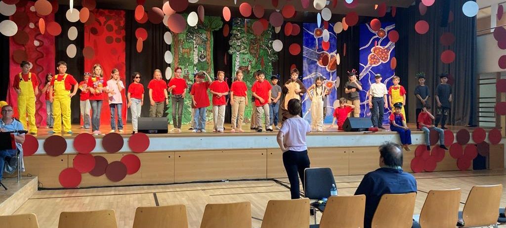 Eine Gruppe von Kindern steht auf einer Bühne mit einem Hintergrund. Einige tragen Kostüme, andere rote Hemden. Eine Frau steht vor ihnen. Das Publikum sitzt in Holzstühlen. Die Bühne hat farbige Kreise, die von der Decke hängen.