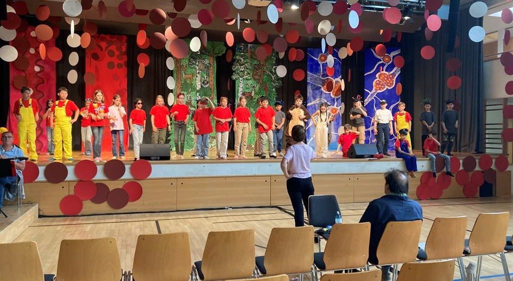 Eine Gruppe von Kindern steht auf einer Bühne mit einem Hintergrund. Einige tragen Kostüme, andere rote Hemden. Eine Frau steht vor ihnen. Das Publikum sitzt in Holzstühlen. Die Bühne hat farbige Kreise, die von der Decke hängen.