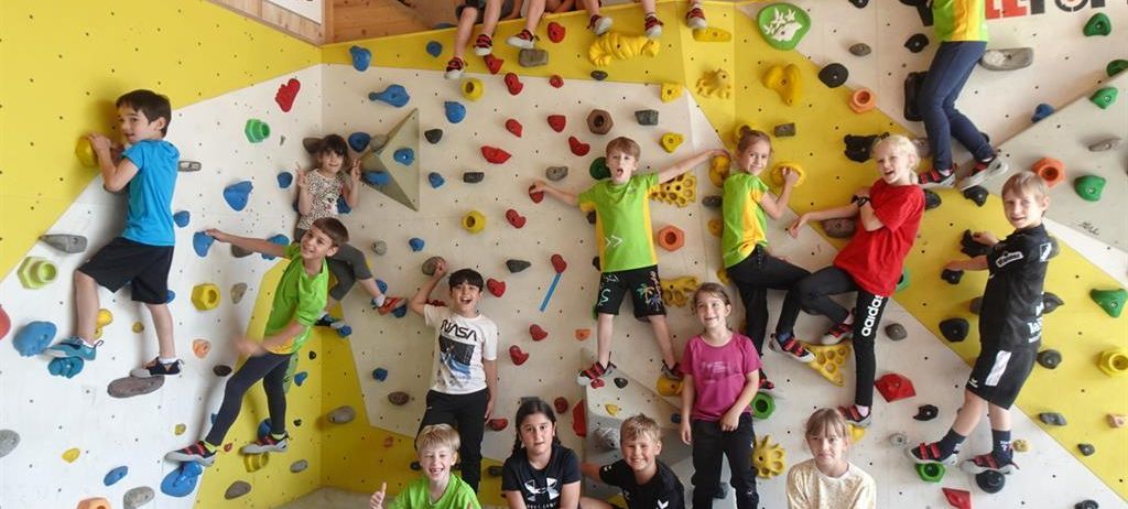 Eine Gruppe von Kindern posiert für ein Foto in einem Kletterzentrum, einige klettern an der Wand, während andere auf dem Boden sitzen. Sie tragen farbenfrohe T-Shirts und Sneakers.