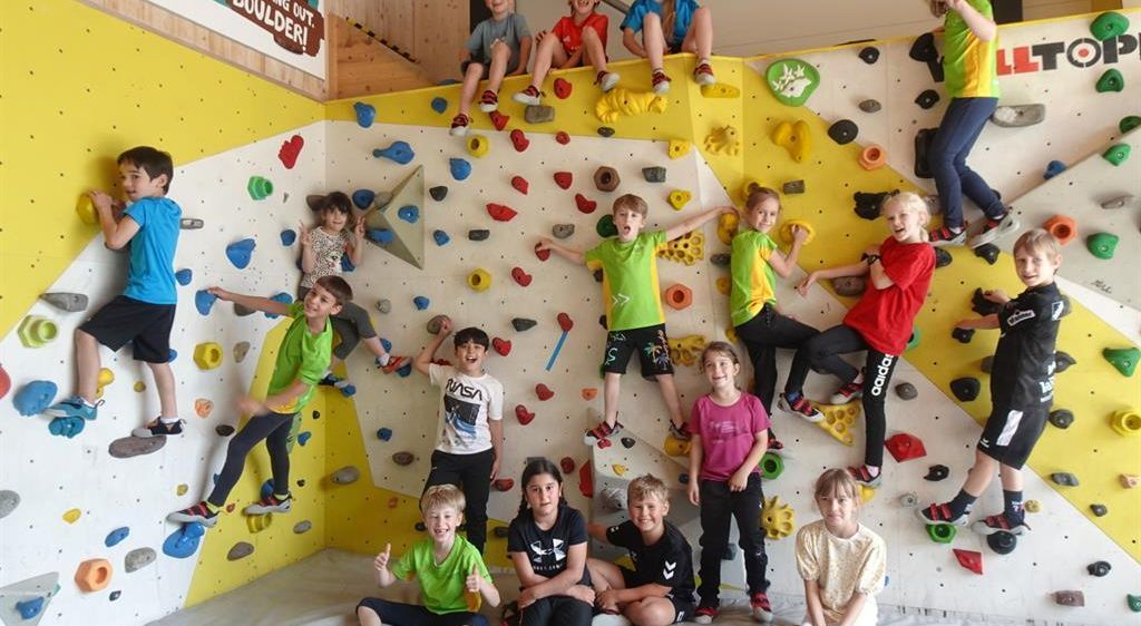 Eine Gruppe von Kindern posiert für ein Foto in einem Kletterzentrum, einige klettern an der Wand, während andere auf dem Boden sitzen. Sie tragen farbenfrohe T-Shirts und Sneakers.