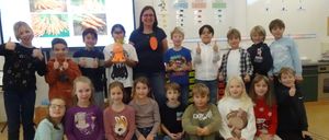 Eine Gruppe von Kindern und einer erwachsenen Frau posiert für ein Foto im Klassenzimmer, einige halten Karotten. Eine Projektionsleinwand im Hintergrund zeigt Karottenbilder.