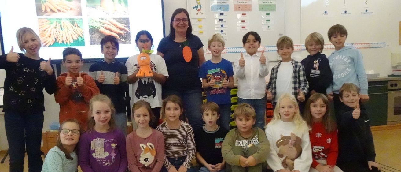 Eine Gruppe von Kindern und einer erwachsenen Frau posiert für ein Foto im Klassenzimmer, einige halten Karotten. Eine Projektionsleinwand im Hintergrund zeigt Karottenbilder.