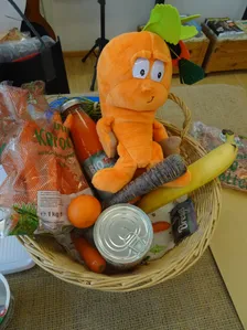 Ein Korb enthält eine orangefarbene Plüschfigur, eine Flasche Saft, einen Beutel Karotten, eine Dose, eine Banane und eine Orange.