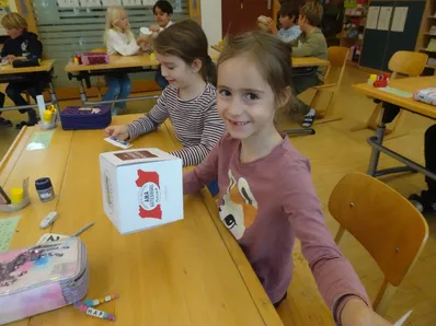 Ein junges Mädchen hält eine weiße Schachtel mit rotem Text und lächelt in einem Klassenzimmer. Andere Kinder sitzen um sie herum an Tischen.