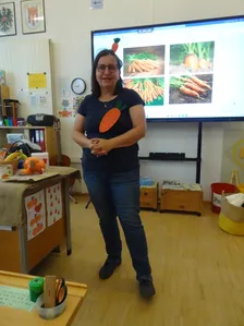 Eine Frau mit einem Karotten-Kopfband steht in einem Klassenzimmer mit einer Projektionsleinwand hinter ihr, die Bilder von Karotten zeigt.