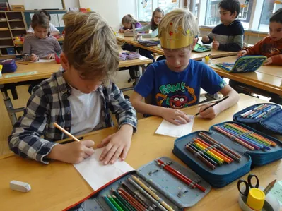 In einem Klassenzimmer sitzen zwei Jungen an Schreibtischen und konzentrieren sich auf das Ausmalen. Der Junge links trägt ein kariertes Hemd und hält einen Bleistift. Der Junge rechts trägt ein blaues T-Shirt, eine goldene Krone und hält einen Bleistift und ein Blatt Papier.