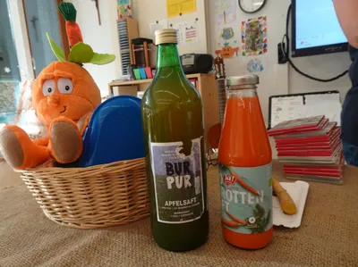 Eine grüne Flasche mit dem Aufkleber 'Bur Pur' steht neben einer Orangensaftflasche mit Karottenaufkleber. Ein Korb aus Weide enthält ein Kuscheltier-Karotte und einen blauen Teller.