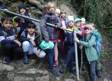 Eine Gruppe von Kindern posiert für ein Foto im Freien. Sie stehen auf felsigem Gelände mit einem Metallgeländer. Einige tragen Mützen, Brillen und Stiefel. Die Umgebung ist grün, mit Pflanzen und Felsen.