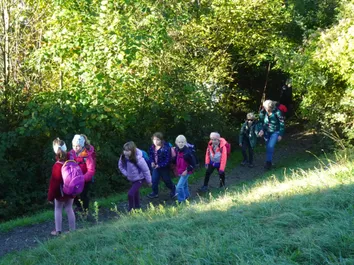 Eine Gruppe junger Mädchen und zwei Erwachsene gehen in einem Wald, tragen Jacken und Rucksäcke. Sie sind auf einem schmalen Pfad umgeben von grünen Bäumen.