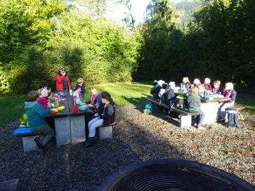 Eine Gruppe von Menschen sitzt um Picknicktische in einem Außenbereich. Einige essen, andere unterhalten sich. Bäume und Berge sind im Hintergrund zu sehen.