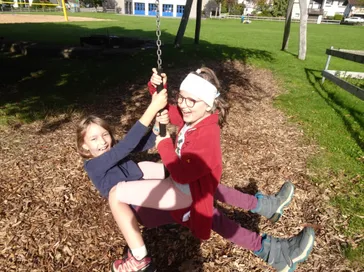 Zwei junge Mädchen spielen auf einer Schaukel in einem Park, eine trägt eine Brille und eine rote Jacke.