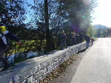 Eine Gruppe von Kindern in Jacken und Turnschuhen geht entlang einer Steinmauer in der Nähe eines Flusses. Sie befinden sich möglicherweise auf einer Klassenfahrt.