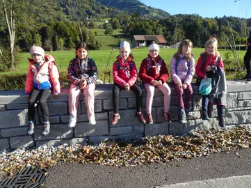 Sechs Mädchen sitzen auf einer Steinmauer. Sie tragen Winterkleidung. Hinter ihnen ist ein Feld und ein Haus. Der Himmel ist klar.
