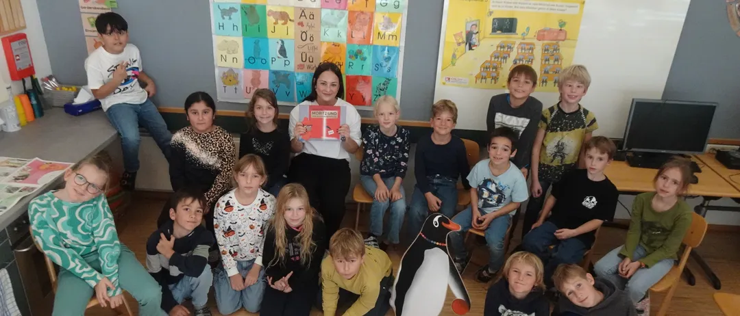 Eine Gruppe von Kindern und einer Frau posiert für ein Foto in einem Klassenzimmer mit einer bunten Alphabetwand und einem Pinguin-Maskottchen.
