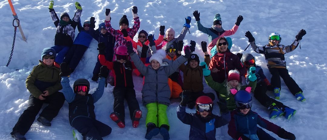 Eine Gruppe junger Menschen posiert für ein Foto auf einem verschneiten Hügel. Sie tragen Winterkleidung und Skiausrüstung, einige heben die Hände. Ein Skistock und Ketten sind sichtbar.