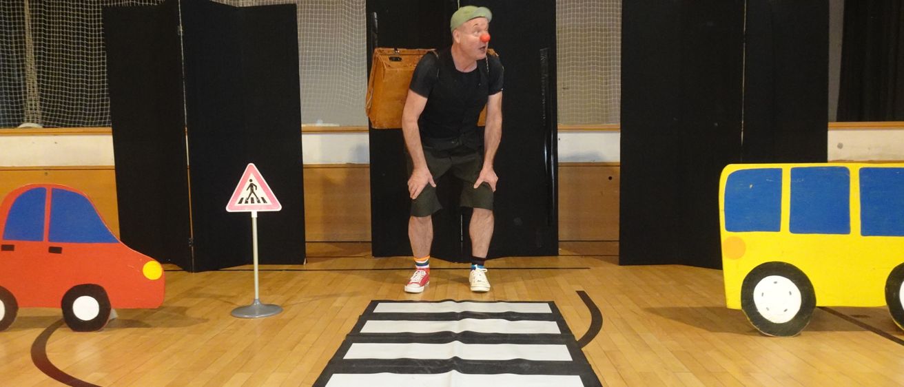 Ein Mann in Clownkostüm steht auf einem gemalten Zebrastreifen, trägt eine Tasche auf dem Rücken, in einer simulierten Bühnenaufstellung mit einem roten Auto und einem gelben Bus.