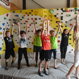Eine Gruppe von Kindern in Sportbekleidung hebt in einem Raum mit Kletterwand die Hände.