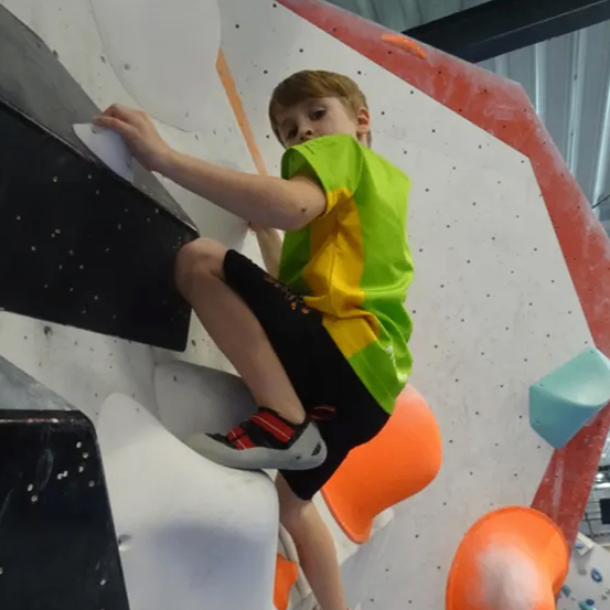 Ein junger Junge klettert eine Kletterwand mit verschiedenen farbigen Griffen. Er trägt ein grünes Shirt und schwarze Shorts. Die Wand ist weiß gestrichen mit roten und orangefarbenen Akzenten.