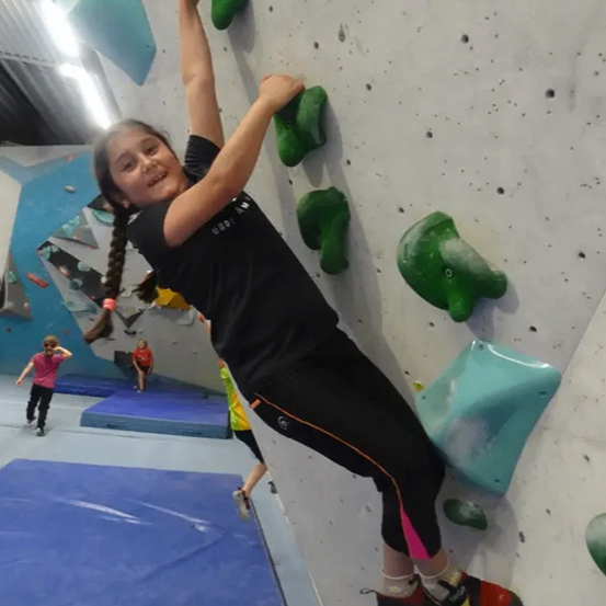 Ein junges Mädchen klettert in einem Fitnessstudio an einer Kletterwand und trägt ein schwarzes Shirt und schwarze Hose. Im Hintergrund sind andere Kletterer zu sehen.