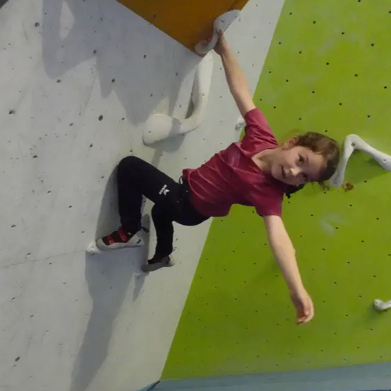 Ein junges Mädchen klettert an einer Felswand und trägt ein rotes T-Shirt und schwarze Hose. Sie benutzt ihre Hände und Füße, um die Wand zu greifen, und ihr Schatten ist an der Wand sichtbar.