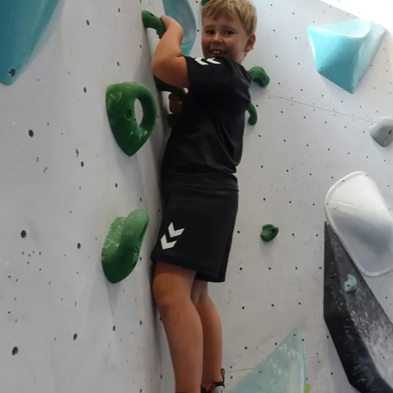 Ein Junge in einem schwarzen T-Shirt und Shorts klettert an einer Kletterwand mit verschiedenen Griffen und hält sich an einem grünen Griff fest, während er lächelt.