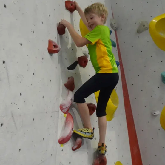 Ein junger Junge klettert an einer Wand mit verschiedenen Griffen. Er trägt ein grün-gelbes Shirt, schwarze Hose und Turnschuhe. Die Wand hat farbige Griffe.