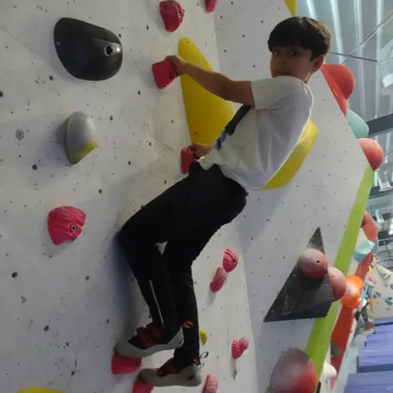 Ein junger Junge klettert an einer farbenfrohen Kletterwand mit verschiedenen Griffen und Haltegriffen. Er trägt ein weißes T-Shirt und schwarze Hose.