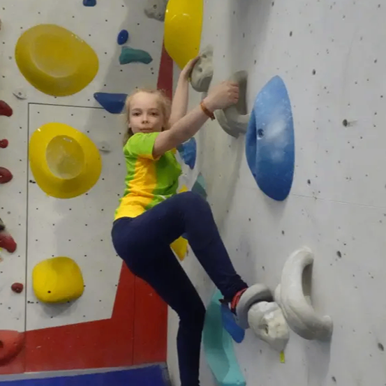 Ein junges Mädchen klettert an einer Kletterwand. Sie trägt ein grün-gelbes Shirt, blaue Hose und Turnschuhe. Die Wand hat verschiedene farbige Griffe.