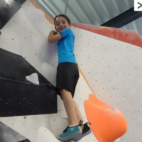 Ein junger Junge klettert an einer Felswand und trägt ein blaues T-Shirt und schwarze Shorts. Er lächelt und scheint die Erfahrung zu genießen.