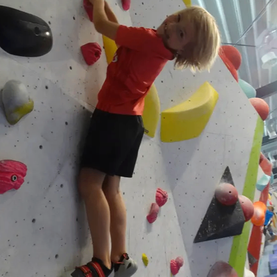 Ein junger Junge klettert an einer Kletterwand mit verschiedenen Griffen. Er trägt ein rotes T-Shirt und schwarze Shorts. Die Wand ist mit bunten Formen verziert.