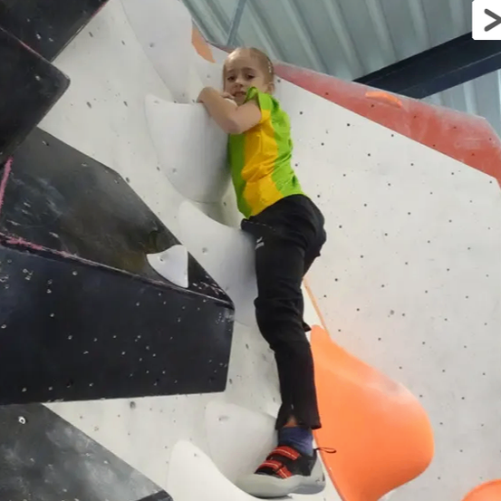 Ein junges Kind in einem grün-gelben Shirt klettert an einer Indoor-Kletterwand mit schwarzen und weißen Griffen.