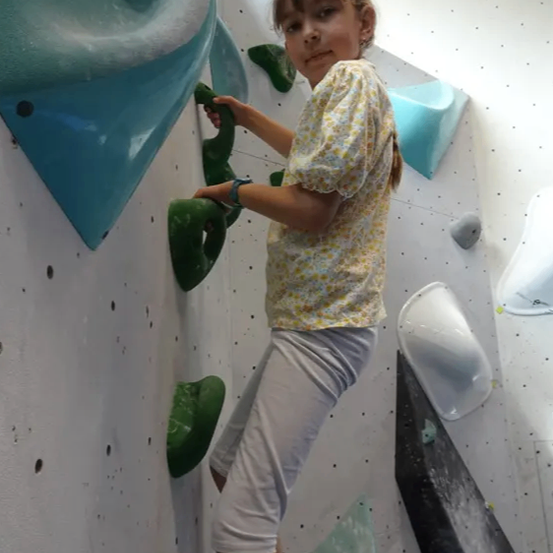 Ein junges Mädchen klettert an einer Kletterwand. Sie hält sich mit der rechten Hand an einem grünen Griff fest. Sie trägt ein Blumenhemd und weiße Hosen. Die Wand hat verschiedene Griffe und Haltepunkte, und es steht ein Markenname, Wallto, darauf.