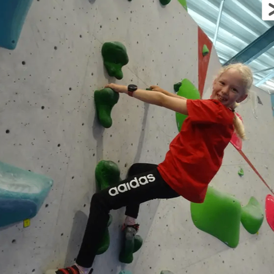Ein junges Mädchen klettert an einer Wand mit bunten Griffen. Sie trägt ein rotes Adidas-Shirt und schwarze Hose. Sie sieht glücklich aus und konzentriert sich auf das Klettern.