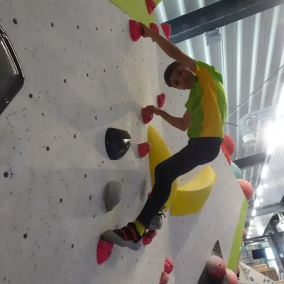 Ein Mann in einem grün-gelben Shirt klettert eine Kletterwand mit verschiedenen farbigen Griffen und Halten. Die Wand hat einen weißen Hintergrund und umfasst grüne und gelbe Abschnitte.