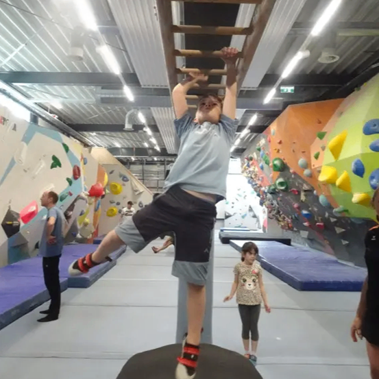 Ein Junge in einem Fitnessstudio hängt von einer Holzleiter. Er trägt Shorts und ein T-Shirt. Ein Mädchen und ein Mann stehen an der Seite. Die Wände haben farbige Steine.