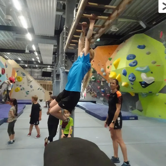 Ein Mädchen klettert in einem Fitnessstudio mit bunten Wänden eine Leiter. Eine Frau steht in der Nähe und beobachtet. Andere Kinder spielen im Hintergrund.