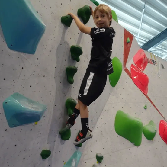Ein junger Junge in einem schwarzen Shirt klettert an einer Felswand mit verschiedenen farbigen Griffen. Er greift einen grünen Griff mit seiner rechten Hand.