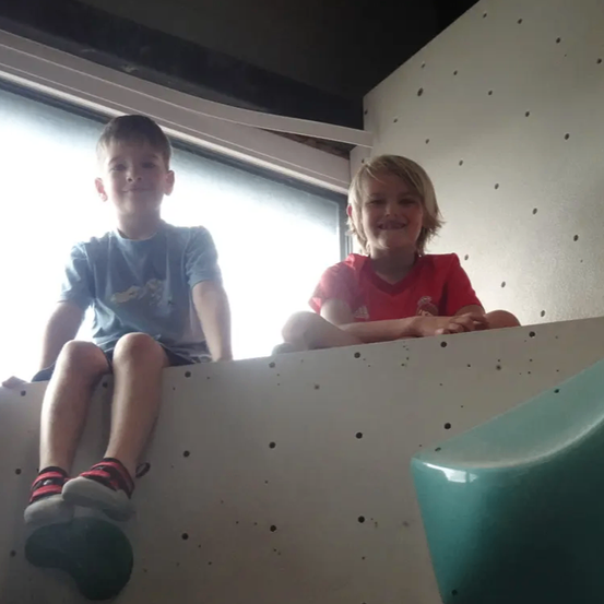 Zwei junge Jungen sitzen auf einer weißen Kletterwand mit einem grünen Objekt davor. Sie lächeln, einer lehnt sich gegen die Wand.