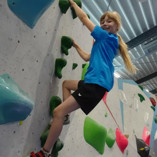 Ein junges Mädchen klettert an einer Wand mit bunten Griffen und trägt ein blaues T-Shirt und schwarze Shorts. Sie lächelt und zeigt ihre Freude.