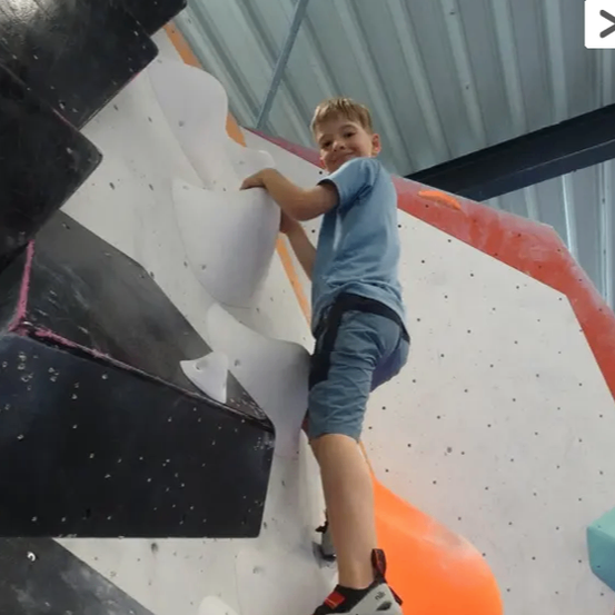 Ein junger Junge klettert an einer Kletterwand, mit dem Fuß auf einem weißen Griff. Er trägt ein blaues T-Shirt und Shorts.