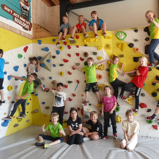 Eine Gruppe von Kindern ist an einer gelben und weißen Kletterwand. Einige klettern, während andere auf der Wand sitzen und stehen. Sie alle lächeln und scheinen für ein Foto zu posieren.